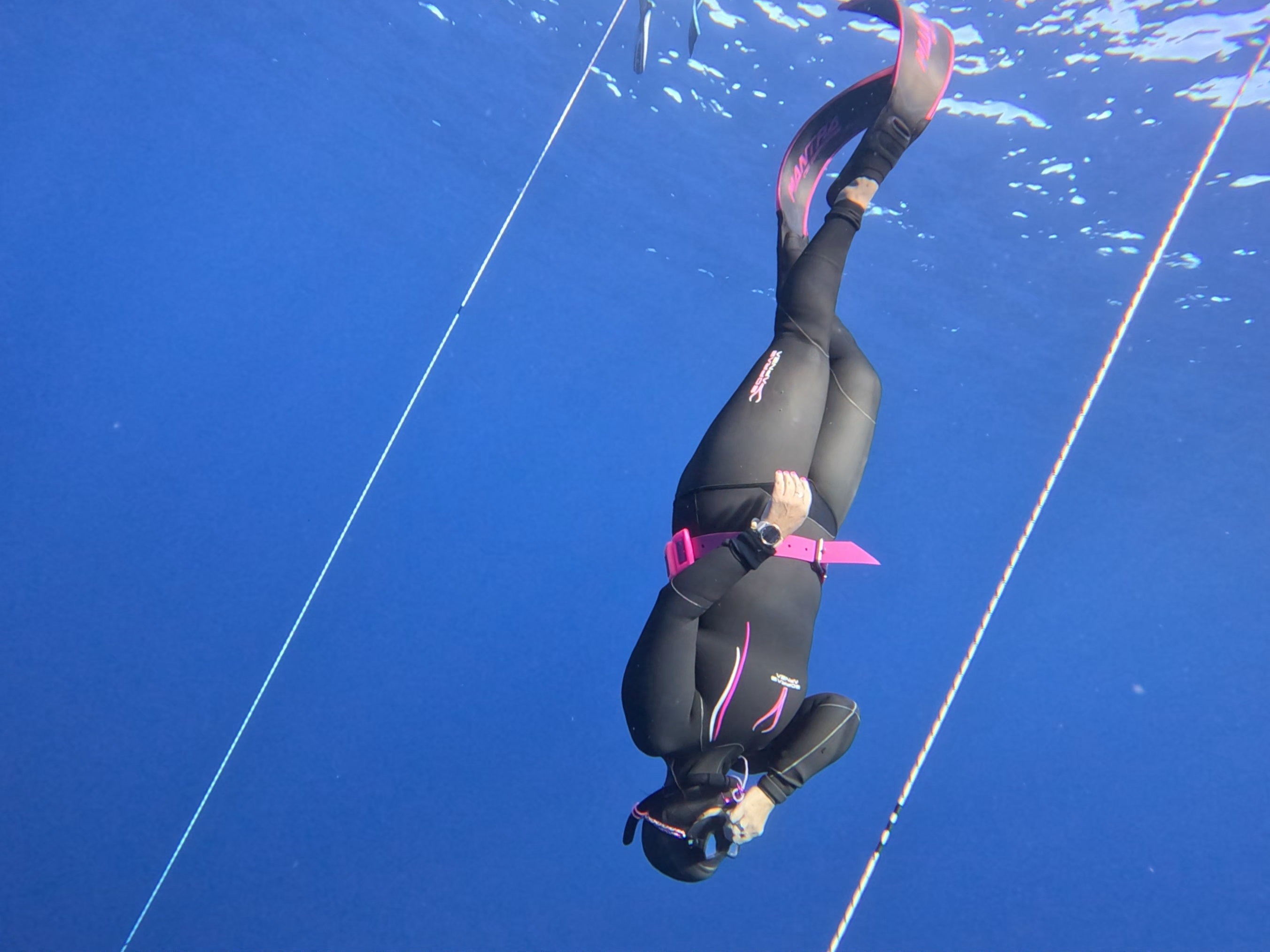 Elaine Hicks descending between two freediving lines in open blue water.